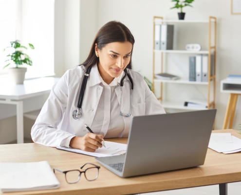 Smiling young friendly female doctor in white uniform sitting at the desk and working with a laptop in medical office